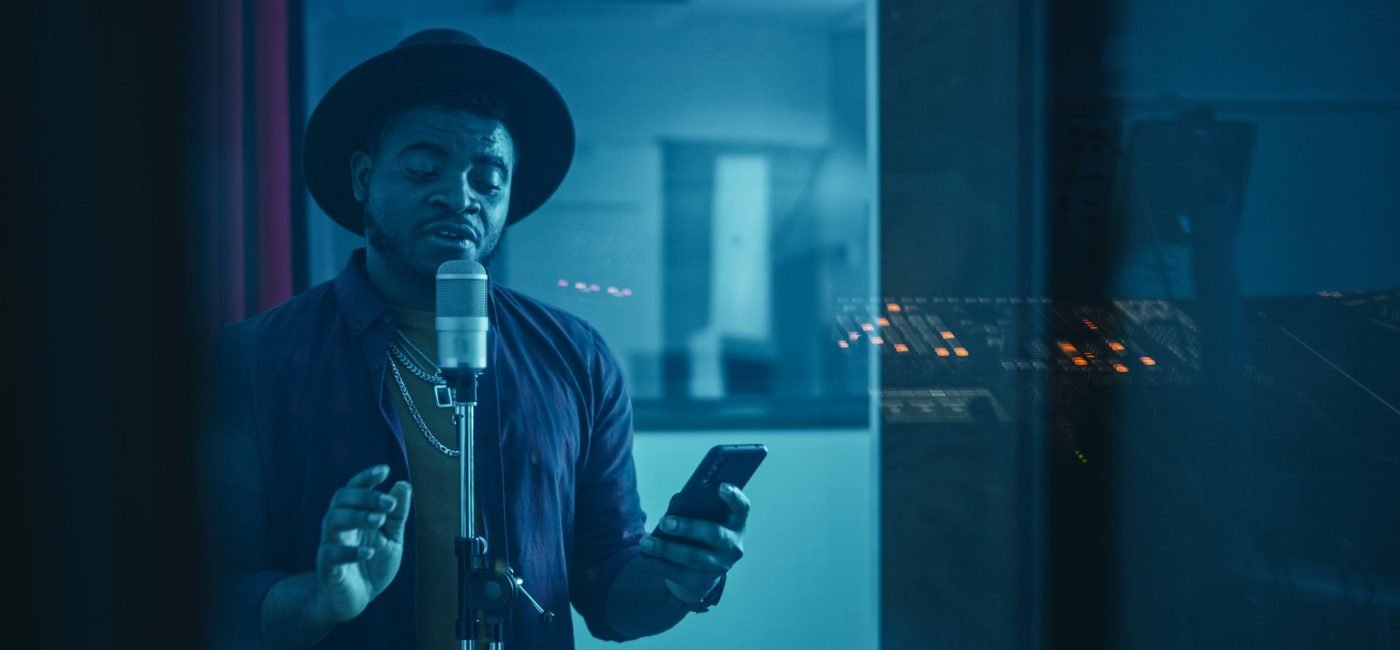 Portrait of Successful Young Black Artist, Singer, Performer Singing His Hit Song for the New Album. Wearing Stylish Hat, Holding Smartphone and Standing in Music Record Studio Soundproof Room.